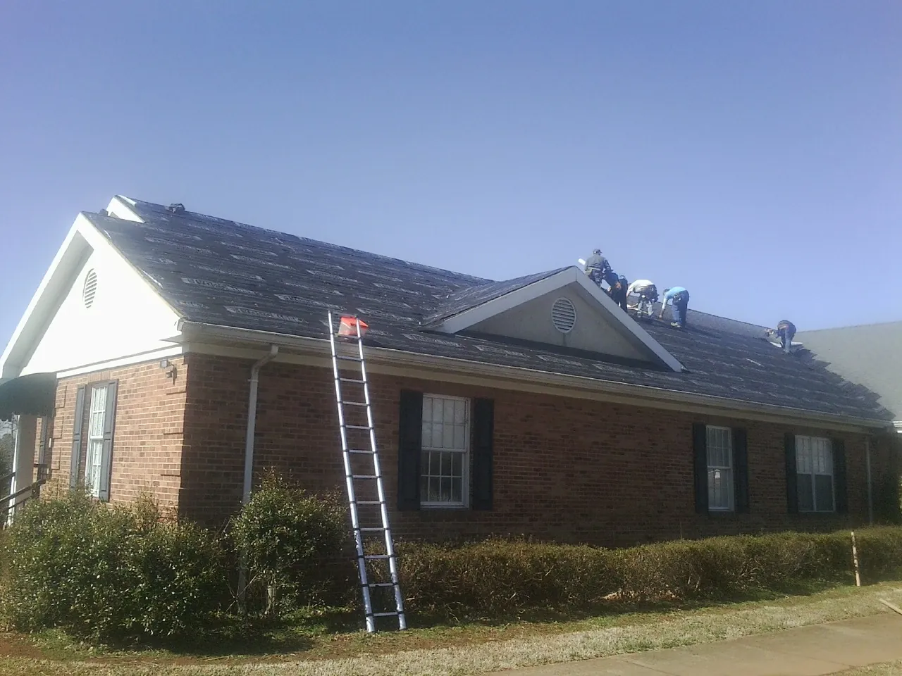 Crew tearing shingles off a brick ranch roof with a ladder set — Metal Roof Repair work by our crew in De Witt
